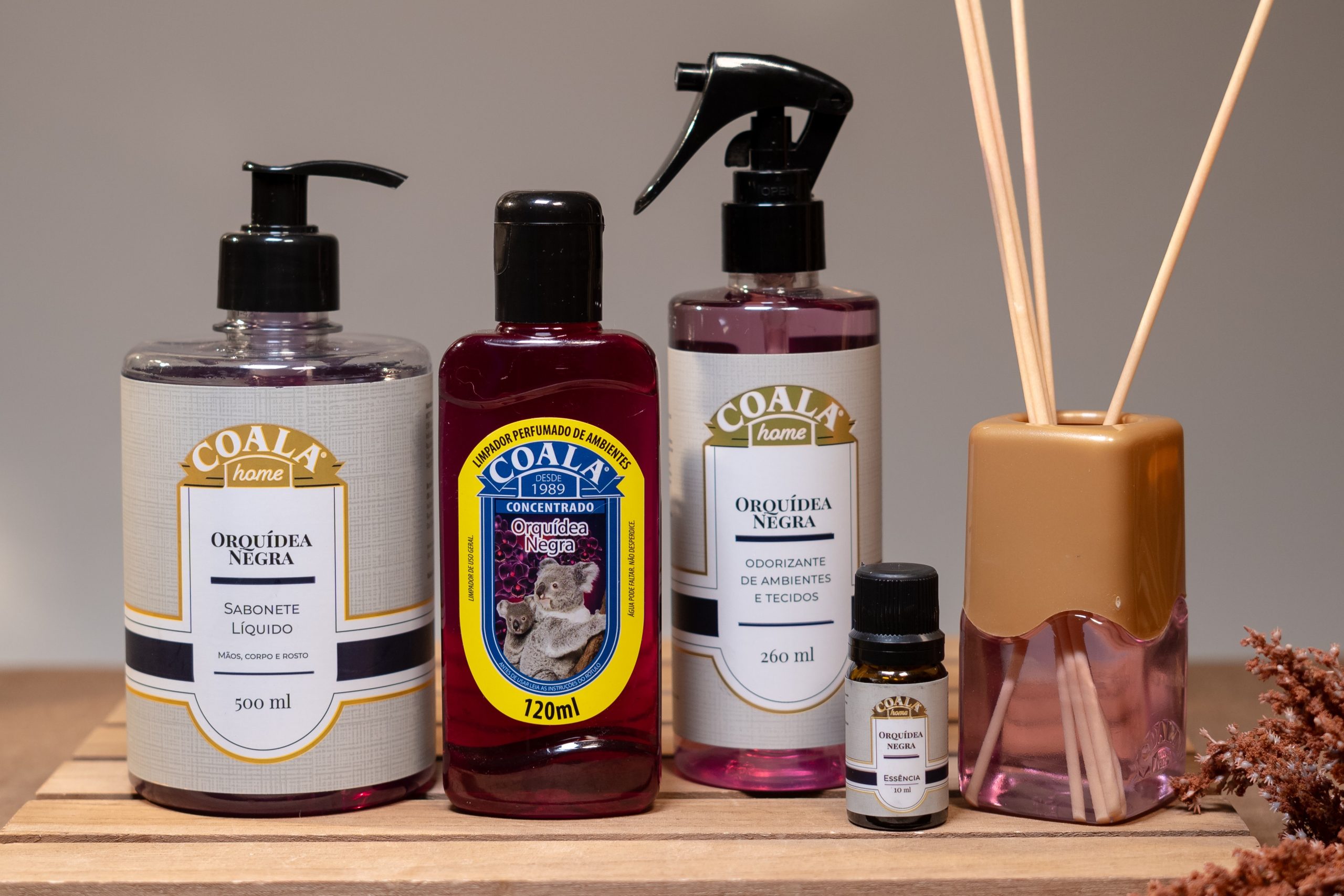 Produtos da linha Coala Orquídea Negra dispostos sobre bancada: sabonete líquido, limpador perfumado concentrado, odorizante de ambientes e tecidos, essência aromática e difusor de aromas.